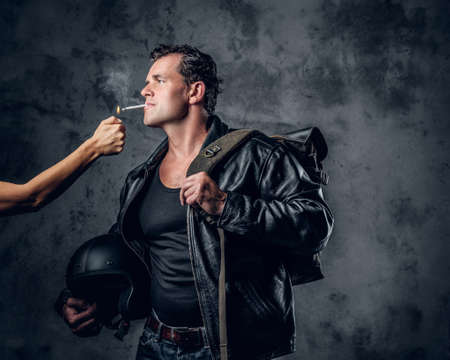Brutal male in leather jacket holds motorcycle helmet and lights up a cigarette from woman arm.の写真素材