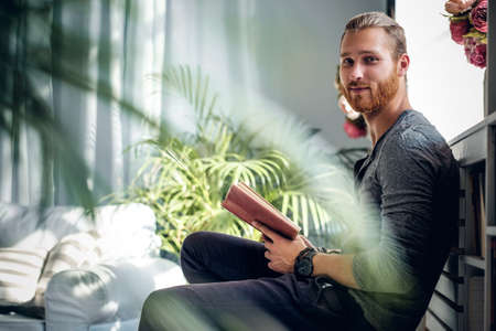 Portrait of young redhead bearded male holding a book in a room with green plants.の写真素材