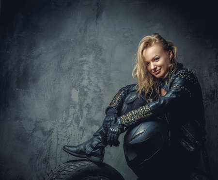 Blonde female in leather clothes sits on a car's wheel and holds safety helmet.の写真素材