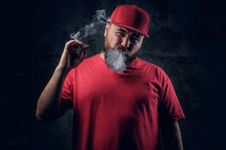 Fatty bearded hipster male in a red hip hop clothes smoking a cigarette on a grey background.の写真素材