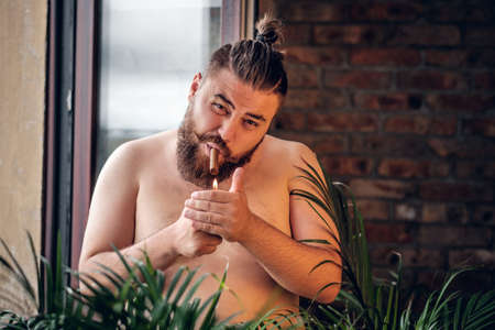 Fatty bearded, shirtless male smoking cigar in front of the window with the wall of the brick on the background.の写真素材