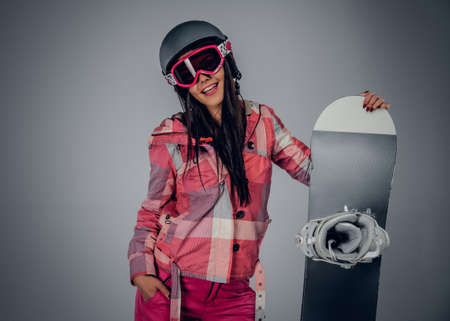 Positive brunette female holding a snowboard on her shoulder in a studio on grey backgroundの写真素材