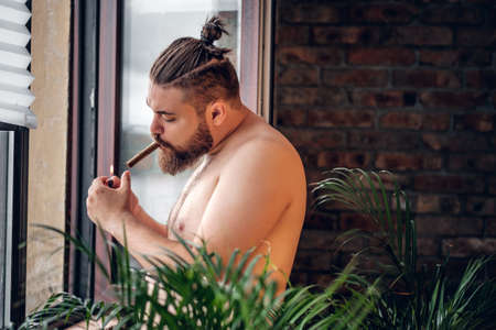 Fatty bearded, shirtless male smoking cigar in front of the window with the wall of the brick on the background.の写真素材