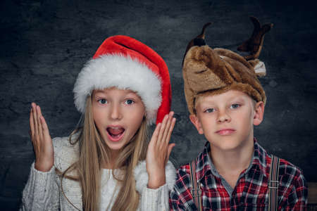 Close up portrait of blonde girl in Santa's hat and cute boy with funny deer hat on grey background.の写真素材
