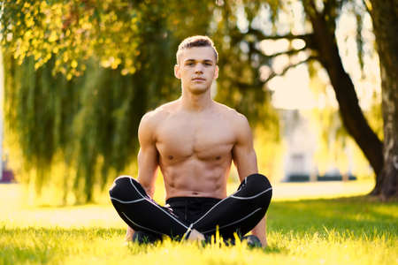 Shirtless abdominal young male fitness model sits on a grass in a park.の写真素材