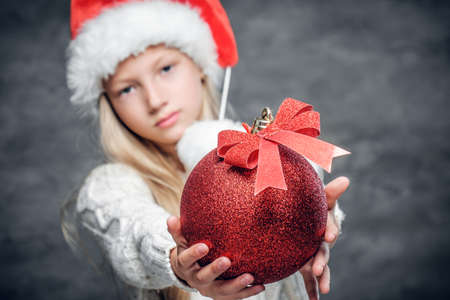 Cute blonde girl in Santa's hat holds red Christmas ball.の写真素材