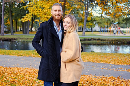 Positive redhead bearded male hugs the cute blonde female near a river in an autumn park.の写真素材