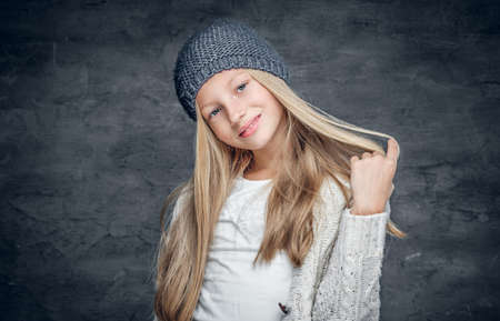 Positive blonde teenage girl in a warm winter hat isolated on grey background.の写真素材