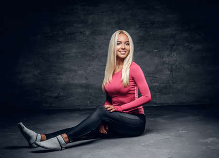 Positive, flexible blonde fitness model sits on a floor in a studio over grey background.の写真素材