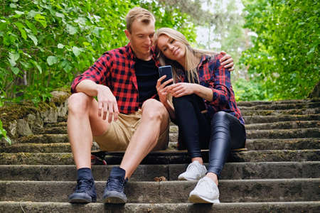 Urban couple relaxing on the steps in wilderness park. A man hugging blonde female using a smartphone.の写真素材
