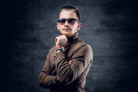 Portrait of blonde, stylish male in sunglasses in turtle-neck sweater on dark grey background.