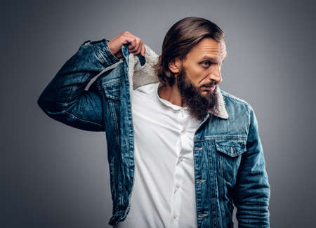 Portrait of bearded male with long hair dressed in a denim jacket, isolated on grey background.の写真素材