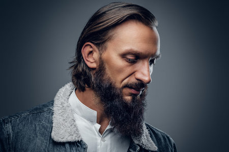 Portrait of bearded male with long hair dressed in a denim jacket, isolated on grey background.