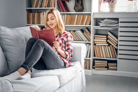 Positive blonde female sits on a couch and reads a book in a living room.の写真素材