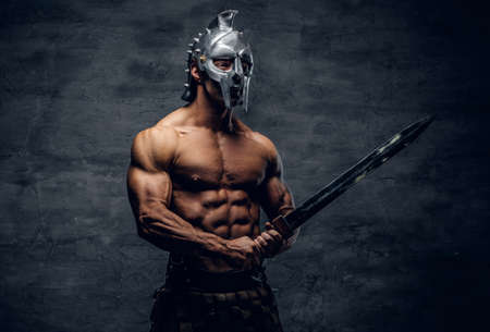 Brutal shirtless muscular male in a silver gladiator helmet holds an iron sword on grey background in a studio.の写真素材