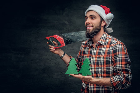 Positive bearded male in Santa's hat holds small fir trees and handsaw on a shoulder.
