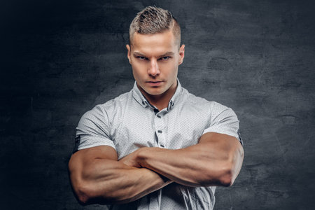 Sporty young male with crossed muscular arms, dressed in a white shirt isolated on grey vignette...