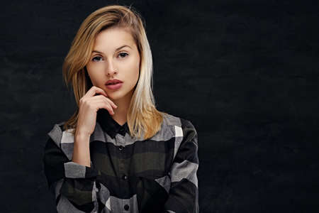 Studio portrait of blonde thoughtful young female dressed in a shirt with square pattern isolated on a dark grey background.の写真素材