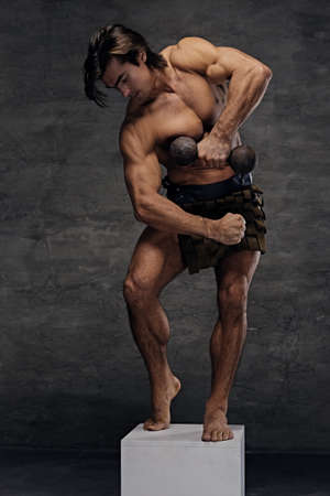 Athletic shirtless male posing on podium and holds dumbbell isolated with contrast illumination on grey background.の写真素材