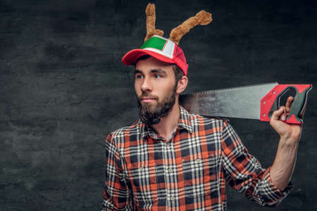 Portrait of bearded male dressed in a plaid shirt and a hat with Christmas deer's horns holds handsaw on a shoulder.の写真素材