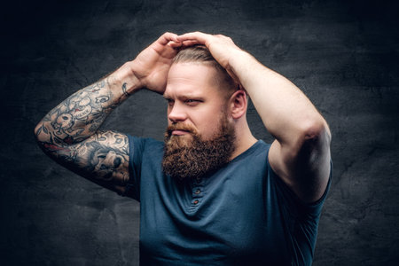 Portrait of bearded male touches his hairstyle with tattooed arms.
