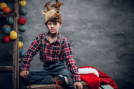 Studio portrait of teenage boy dressed in a plaid shirt and a hat with holiday deer horns on grey background with Christmas colorful lights.の写真素材