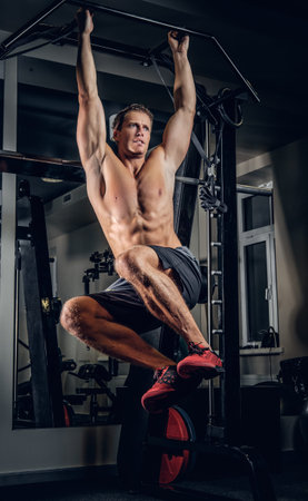 Shirtless sporty male doing ABS workouts on pull up bar in a gym club.
