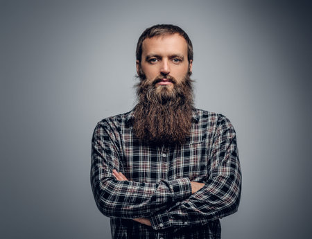 Bearded male with crossed arms on his chest, dressed in a plaid shirt isolated on grey background.
