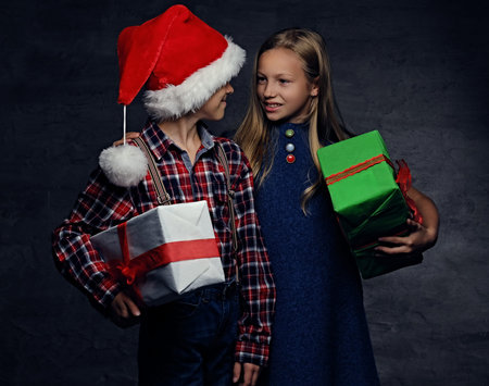 Christmas time. Teenage boy in Santa's hat and blonde girl holds gift boxes on grey background.の写真素材
