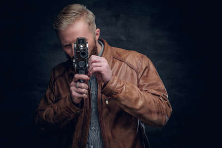 Portrait of blond bearded male dressed in brown leather jacket holds vintage 8mm video camera.の写真素材