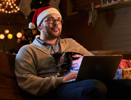 Fatty bearded male in Santa's hat dreaming on a couch with a laptop in a room with Christmas holiday decoration.の写真素材
