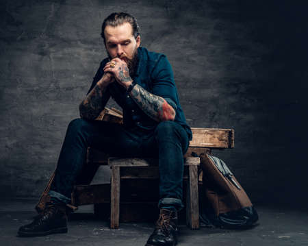 Studio portrait of stylish bearded male with tattoos on arms, sits on wooden boxes over grey background in a studio.の写真素材