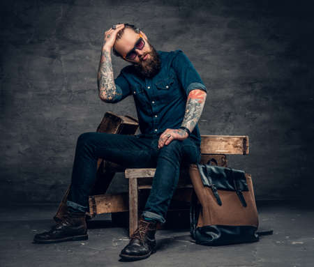 Bearded stylish male in a jeans and sunglasses sits on wooden box over grey background.の写真素材