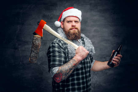 Brutal bearded male in Santa's hat wearing green plaid shirt, drinking beer and holds axe on a shoulder.の写真素材