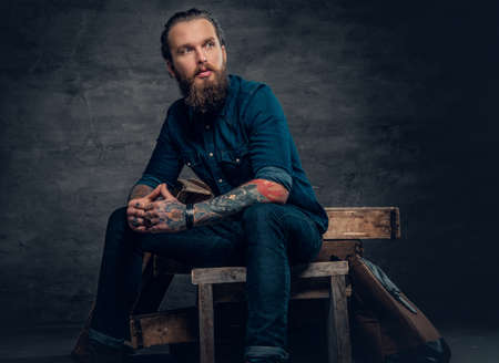 Studio portrait of stylish bearded male with tattoos on arms, sits on wooden boxes over grey background in a studio.の写真素材