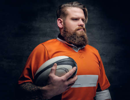 Studio portrait of the bearded rugby player wearing an orange t shirt holds game ball.の写真素材