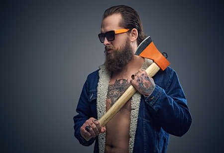 Portrait of bearded male in sunglasses with a tattoo on his chest wearing a denim jacket and holds axe over grey background.の写真素材