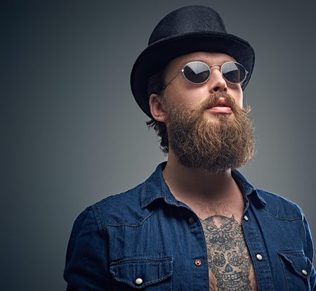 Studio portrait of stylish bearded male with a tattoo on his chest, dressed in a denim shirt,...