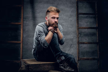 Studio portrait of bearded urban male in a jeans sits on a wooden box with vintage ladders on background.の写真素材