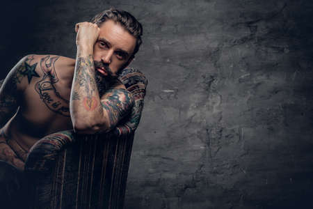 Portrait of bearded male with tattooed naked torso sits on a chair over dark grey background.の写真素材