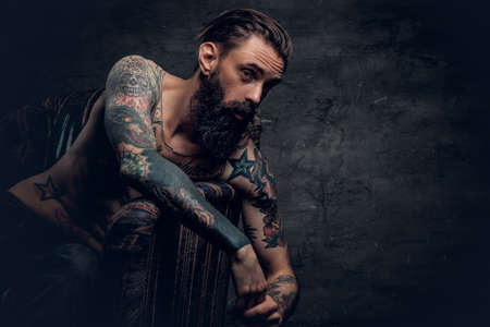 Portrait of bearded male with tattooed naked torso sits on a chair over dark grey background.の写真素材