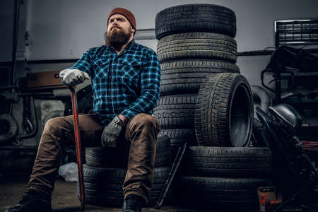 Bearded mechanic sits on an old car's tire in a garage.の写真素材