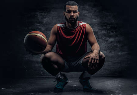 Full body portrait of a black basketball player sits on a floor and holds basket ball.