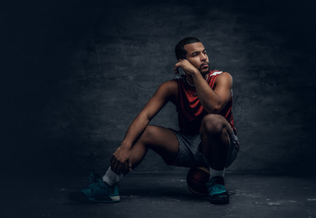 Full body portrait of a black basketball player sits on a floor and holds basket ball.