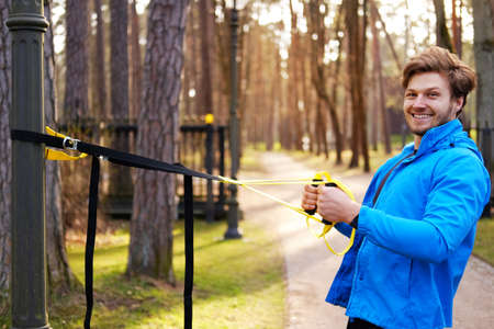 Attractive positive male in a blue raincoat exercising in a park with trx fitness strips.の写真素材