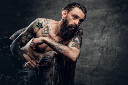 The shirtless, tattooed bearded male sits on a chair over grey background.