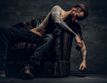 The shirtless, tattooed bearded male sits on a chair over grey background.の写真素材