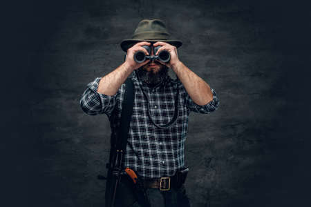 Studio portrait of bearded hunter male wearing a plaid fleece shirt looking through binoculars, holds a rifle.の写真素材