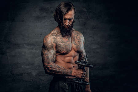 Shirtless bearded male with tattooed body holds dumbbell on grey background.の写真素材