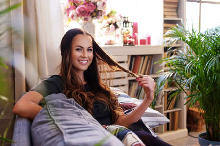 Portrait of a smiling brunette woman sits on a sofa in a room with green palm plants and book stands.の写真素材